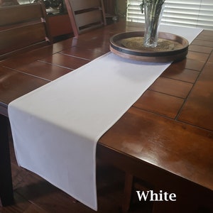 May include: White table runner on a wooden table with a vase of flowers and a wooden tray. The table runner is 14 inches wide and 70 inches long. White.