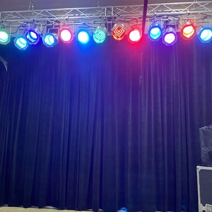 May include: A row of stage lights with different colored lenses, including red, blue, green, and white, are mounted on a metal truss above a black curtain backdrop.