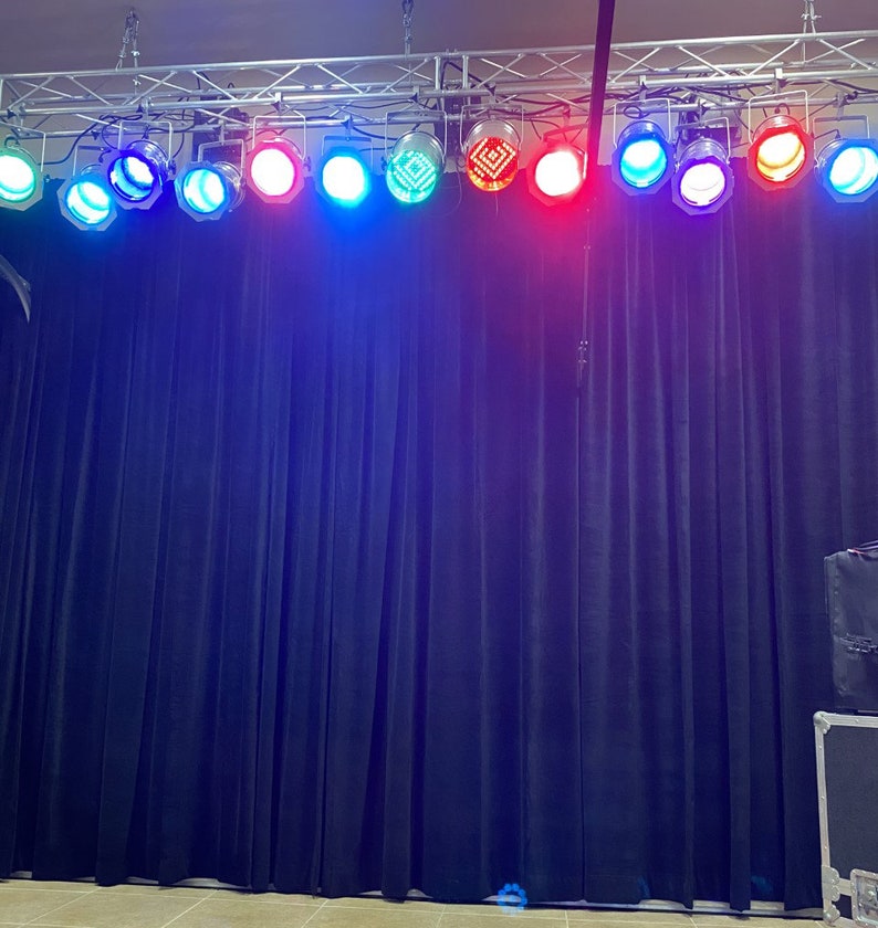 May include: A row of stage lights with different colored lenses, including red, blue, green, and white, are mounted on a metal truss above a black curtain backdrop.