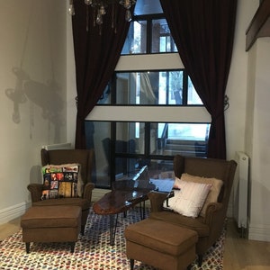 May include: Two brown leather armchairs with matching ottomans, a wooden coffee table with black hairpin legs, and a colorful patterned rug. The chairs are positioned in front of a large window with burgundy curtains.