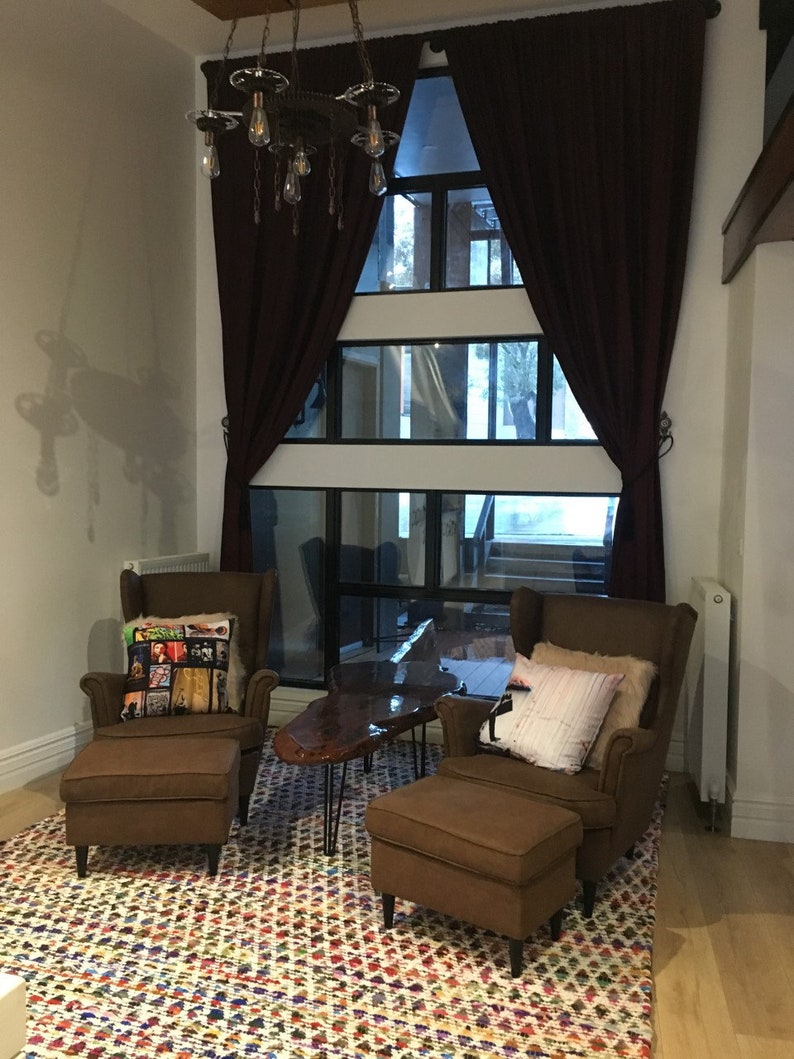 May include: Two brown leather armchairs with matching ottomans, a wooden coffee table with black hairpin legs, and a colorful patterned rug. The chairs are positioned in front of a large window with burgundy curtains.