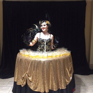 May include: A woman in a black and gold sequined dress and a feather headdress stands behind a table covered in a gold sequined tablecloth. The table is filled with small clear cups filled with a light yellow liquid.