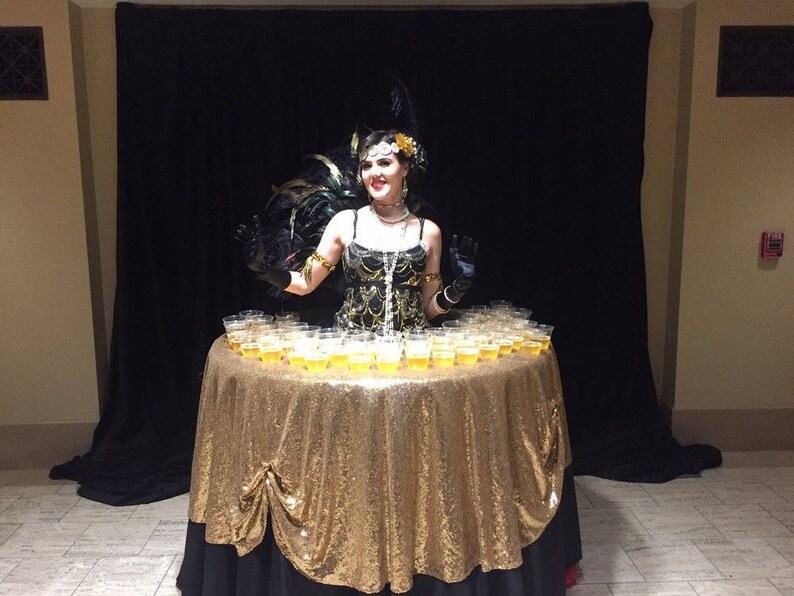 May include: A woman in a black and gold sequined dress and a feather headdress stands behind a table covered in a gold sequined tablecloth. The table is filled with small clear cups filled with a light yellow liquid.