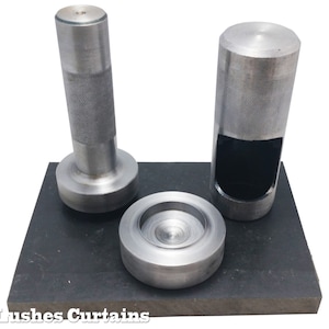 May include: Three metal tools, likely for crafting or industrial use, are arranged on a dark gray rectangular base. The tools include a cylindrical punch with a hollow center, a solid cylindrical tool, and a circular piece. The tools have textured handles.