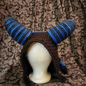 May include: A black and blue crocheted hat with horns. The hat is on a styrofoam head and has a hood.