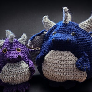 May include: Two crocheted plush dragon toys. The larger dragon is blue and gray, while the smaller dragon is purple and gray.