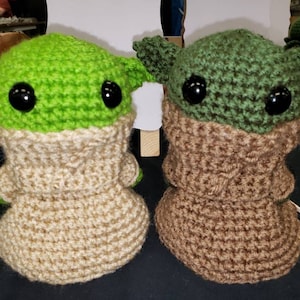 May include: Two crocheted plush toys, one green and one brown, with large black eyes. The green toy has a tan coloured body and the brown toy has a tan coloured body.