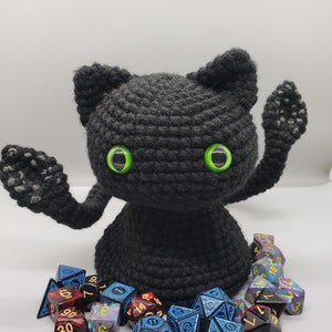 May include: A black crocheted cat toy with green eyes, sitting on a white surface with a variety of colorful dice. The dice are blue, red, and purple with gold numbers.