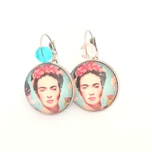 Könnte beinhalten: Ein Paar silberne Ohrringe mit einem runden Glasanhänger, der ein Porträt von Frida Kahlo zeigt. Ein Ohrring hat eine türkisfarbene Perle und der andere eine klare Perle.