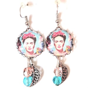 Puede incluir: Un par de pendientes de plata con una imagen redonda de Frida Kahlo sobre un fondo azul. Los pendientes tienen un dije de corazón de plata y una cuenta de cristal azul.