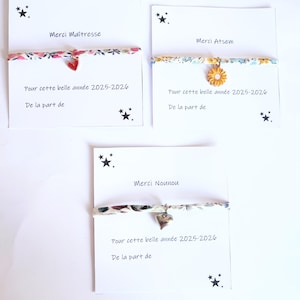 Könnte beinhalten: Drei Stoffarmbänder auf weißen Karten. Eines hat einen roten Herzanhänger, eines einen gelben Sonnenblumenanhänger und eines einen silbernen Herzanhänger. Die Karten zeigen schwarze Sternsymbole und den französischen Text Merci Maîtresse, Merci Atsem, Merci Nounou sowie das Jahr 2025-2026.