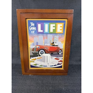 May include: A wooden box containing The Game of Life board game. The front panel features a colorful illustration of a red vintage car driving toward a city skyline, with the text The Game of LIFE displayed prominently above the vehicle.