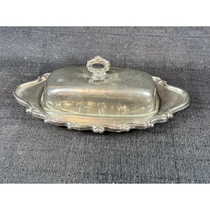 May include: A silver-colored butter dish with a lid and a decorative handle. The dish has an oval shape with a scalloped edge. The lid has a small, ornate handle. The butter dish is likely made of metal.