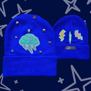 Puede incluir: Dos gorros azul real. Un gorro presenta un parche de medusa con tachuelas doradas. El otro gorro tiene un diseño abstracto colorido y una pequeña etiqueta que dice "Biscuit & Min". El fondo es azul oscuro con formas de estrellas blancas.