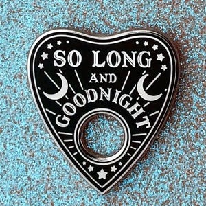 May include: A black and silver heart-shaped planchette with the text "SO LONG AND GOODNIGHT". The planchette features crescent moons, stars, and a circular opening. The design is set against a sparkly, light blue background.