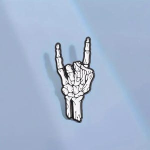 Może przedstawiać: Biała szkieletowa dłoń wykonująca gest rock and roll. Dłoń jest obwiedziona czernią i jest metalową szpilką.