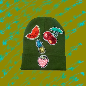 Può includere: Un berretto verde con toppe di frutta ricamate, tra cui un'anguria, ciliegie, ananas e fragola.