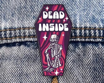 May include: A black and white enamel pin shaped like a coffin with a skeleton inside. The coffin is red and has the words "Dead Inside" written on it.