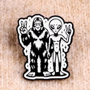 Peut inclure: Épingle en émail noir et blanc représentant un Bigfoot et un extraterrestre, tous deux faisant le signe de la paix. L'épingle a une bordure noire avec des accents blancs, parfaite pour ajouter une touche de fantaisie.