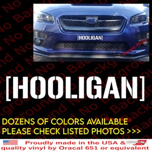 May include: A white vinyl decal with the word "HOOLIGAN" in bold, block letters. The decal is shown on the front bumper of a blue car. The text "DOZENS OF COLORS AVAILABLE. PLEASE CHECK LISTED PHOTOS >>" is shown below the decal.