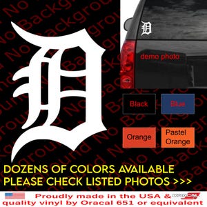 May include: White vinyl decal of a stylized letter D, with a black background. The decal is shown on a car window. The text "DOZENS OF COLORS AVAILABLE PLEASE CHECK LISTED PHOTOS >>>" is at the bottom of the image.