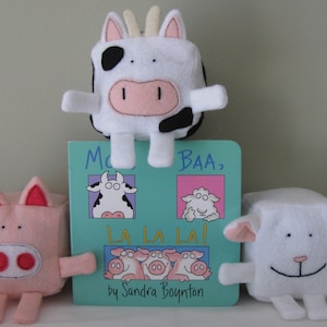 May include: A children's book titled "Moo, Baa, La La La!" by Sandra Boynton, featuring illustrations of a cow, sheep, and pig. The book is displayed with three plush toys: a cow, a sheep, and a pig.