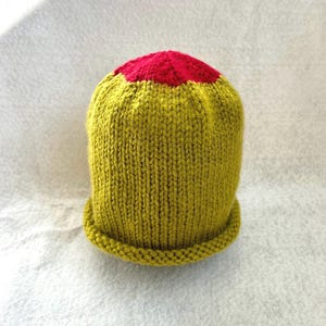 May include: A hand-knitted beanie featuring a vibrant olive green body with a contrasting bright red circular crown. The hat is constructed with a vertical ribbed stitch pattern and includes a rolled brim edge at the base for a finished look.