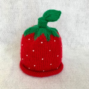 May include: A knitted strawberry hat in red with white bead accents. The hat features a green stem and leaf detail. The hat is designed to look like a strawberry.