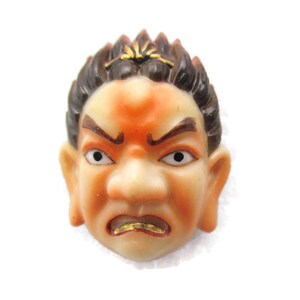 May include: A small ceramic figurine of a man's face with a scowling expression. The figurine is painted in shades of orange, brown, and gold.