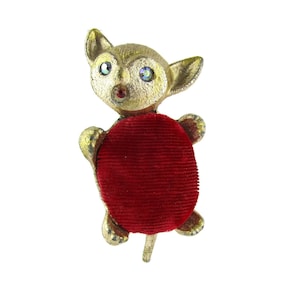 May include: A vintage pin cushion shaped like a gold-toned animal with a red velvet cushion body. The animal has large ears, a small nose, and eyes with iridescent accents. The pin cushion is set against a white background.