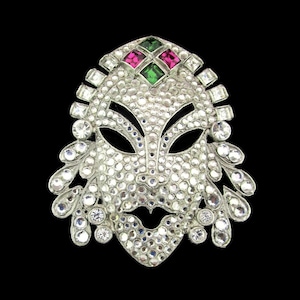 May include: A silver tone mask brooch with rhinestones and green and pink colored stones.