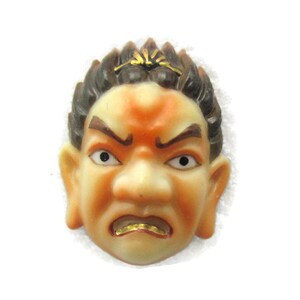 May include: A ceramic mask with a stylised human face. The mask has brown and black hair, a light orange face, and a gold painted mouth. The mask has a frowning expression.