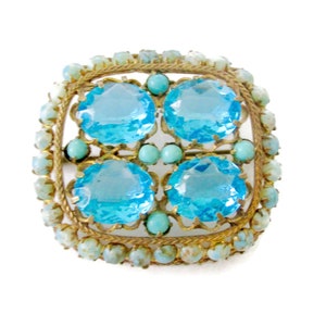 Puede incluir: Un broche dorado con un diseño cuadrado que presenta cuatro piedras preciosas grandes, facetadas, de color azul claro rodeadas de piedras preciosas más pequeñas de color azul claro.