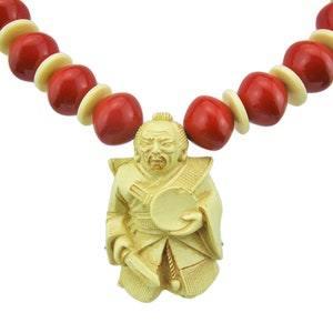 Peut inclure: Un collier avec des perles de couleur rouge et crème et un pendentif en crème représentant une figure sculptée tenant un éventail.