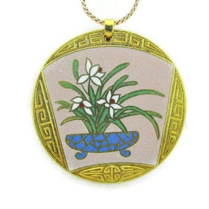Puede incluir: Un collar con un colgante redondo de esmalte rosa y blanco con un diseño floral. El colgante tiene una maceta de esmalte azul y dorado con flores blancas y hojas verdes. El colgante está rodeado por un borde dorado con un patrón geométrico.