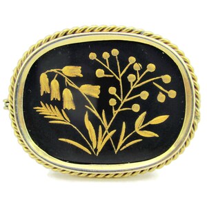 Puede incluir: Un broche dorado con fondo de esmalte negro que presenta un dise&ntilde;o floral detallado en oro. Las flores son estilizadas y tienen un aspecto delicado e intrincado.