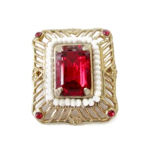 Könnte beinhalten: Eine goldfarbene Brosche mit einem roten Edelstein, umgeben von weißen Perlen und kleineren roten Edelsteinen.