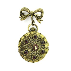 May include: A gold-tone brooch with a round pendant featuring a cameo and red jewels. The pendant is attached to a bow-shaped pin.