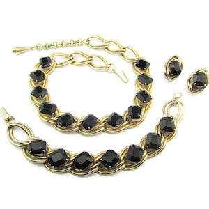 以下が含まれることがあります： 黒いファセットカットの石が付いたゴールドトーンのネックレス、ブレスレット、イヤリングのセット。ジュエリーはチェーンのデザインでつながっています。