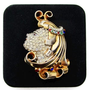 May include: A gold-tone brooch featuring a stylized woman's face with rhinestones and colorful accents.