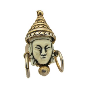 Puede incluir: Un colgante dorado y blanco con un rostro tallado con un sombrero. El colgante está unido a un anillo de plata.