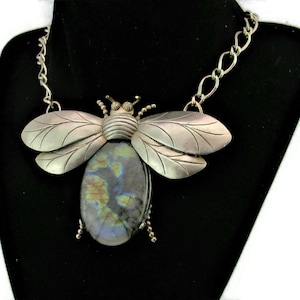 Puede incluir: Un collar de abeja de plata con una gran piedra preciosa azul y verde iridiscente. La abeja tiene alas detalladas y un cuerpo texturizado.