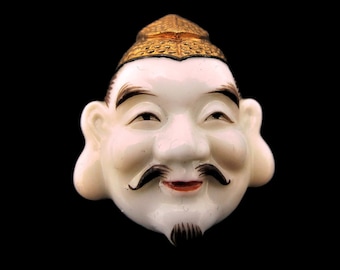 May include: A vintage, off-white ceramic mask of a smiling Asian man with black facial hair and a gold-colored hat. The mask has a serene expression and is set against a black background.