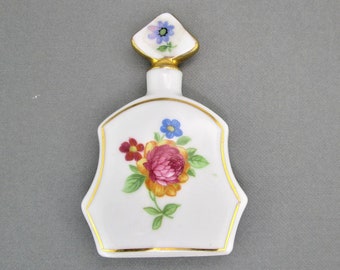 Könnte beinhalten: Eine weiße Porzellan-Parfümflasche mit goldener Verzierung und floralem Design. Die Flasche hat einen rautenförmigen Stopfen mit einer darauf gemalten blauen Blume.