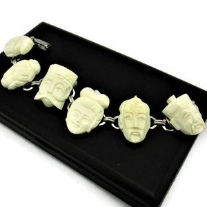 May include: A white bone bracelet with carved faces of people. The bracelet is on a black velvet background.