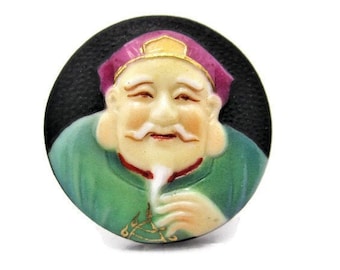 May include: A round ceramic button with a raised image of a smiling man with a long white beard wearing a green robe and a pink and gold hat. The button has a black background.