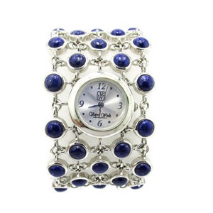 Könnte beinhalten: Eine silberne Uhr mit weißem Zifferblatt und blauen Edelstein-Akzenten. Die Uhr hat ein einzigartiges Armband im Manschetten-Stil.