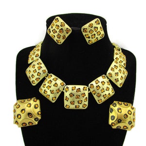 Op de afbeelding: Een gouden ketting- en oorsteensets met een geometrisch ontwerp. De ketting is gemaakt van meerdere vierkante stukken, elk versierd met kleine, glinsterende amberkleurige strass steentjes. De oorbellen zijn bijpassende vierkante stukken.