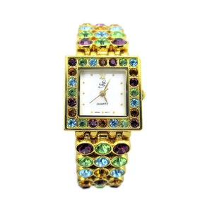 May include: A gold-tone watch with a white face and a crystal-encrusted band. The band is made up of small, colorful stones in shades of green, blue, purple, and orange. The watch face has black Roman numeral hour markers and the words "QUARTZ" and "Ella" printed on it.