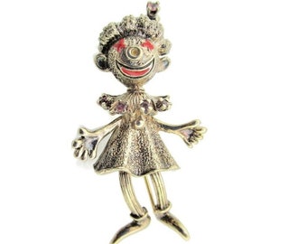 May include: Gold-toned pin of a smiling clown doll with red and pink accents. The clown has a red nose, red eyes, and a pink hat. The doll is wearing a pink and white dress and has long, thin legs.